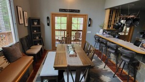 Un comedor con una larga mesa de madera, sillas y un banco. Se observa una zona de cocina con ollas colgadas y un letrero sobre la puerta.