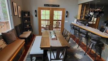 A dining area with a long wooden table, chairs, and a bench. There is a kitchen area with hanging pots and a sign above the door.