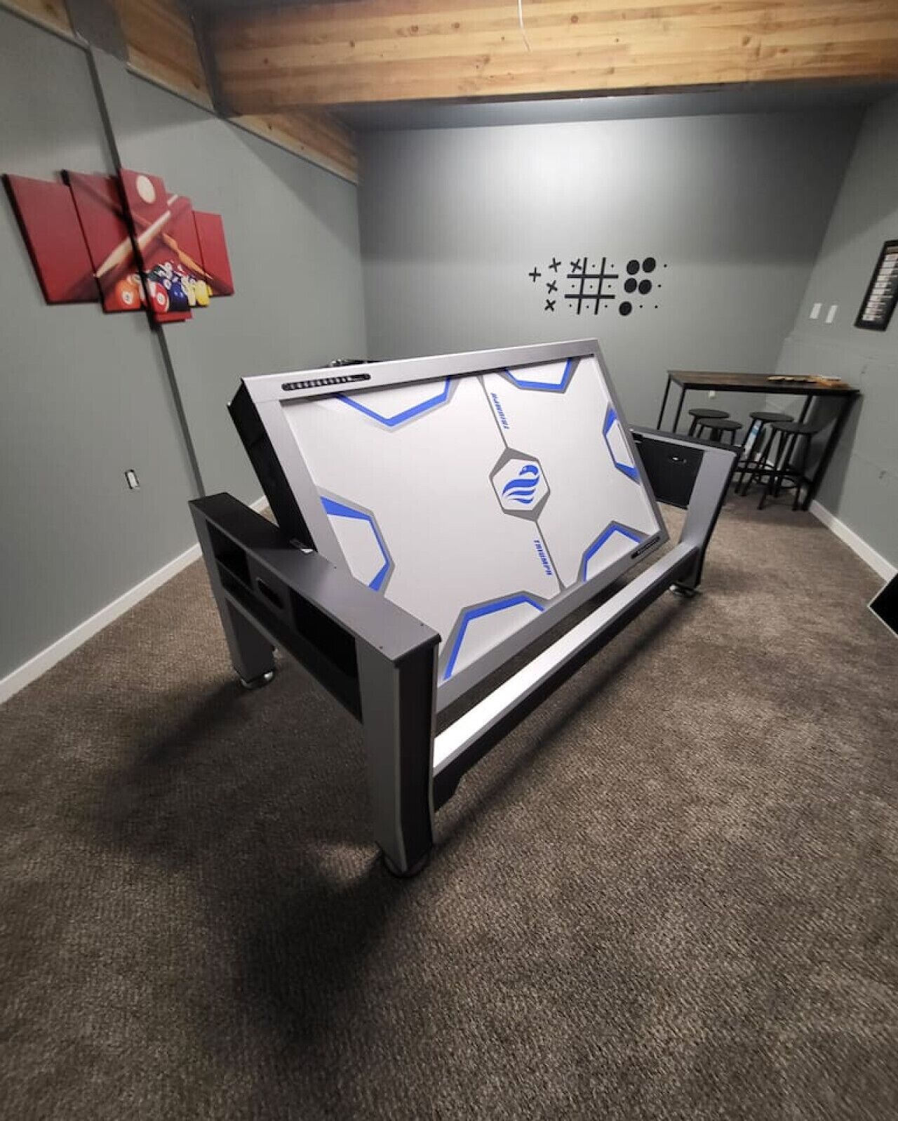 Une pièce dotée d’une grande table de jeux, de murs gris, d’un plafond en bois et d’un petit coin bar.
