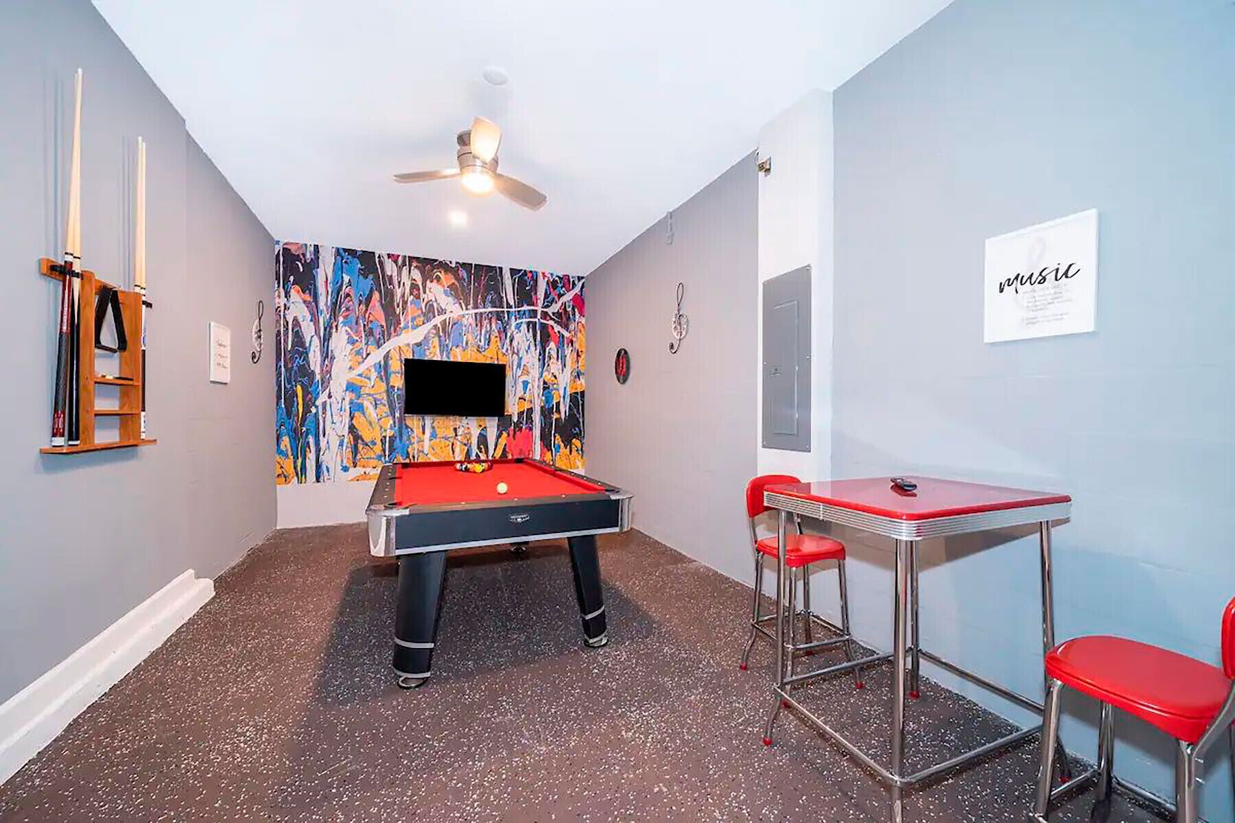 Ein Billardtisch mit roter Spielbahndecke, zwei rote Sessel, ein Deckenventilator und ein farbenfrohes, abstraktes Wandbild.