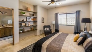 Um quarto com cama, uma cadeira, escrivaninha, prateleiras com objetos decorativos, janela com cortinas e um ventilador de teto.