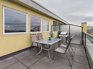 A balcony with a table and chairs, a grill, and large windows overlooking a cityscape.