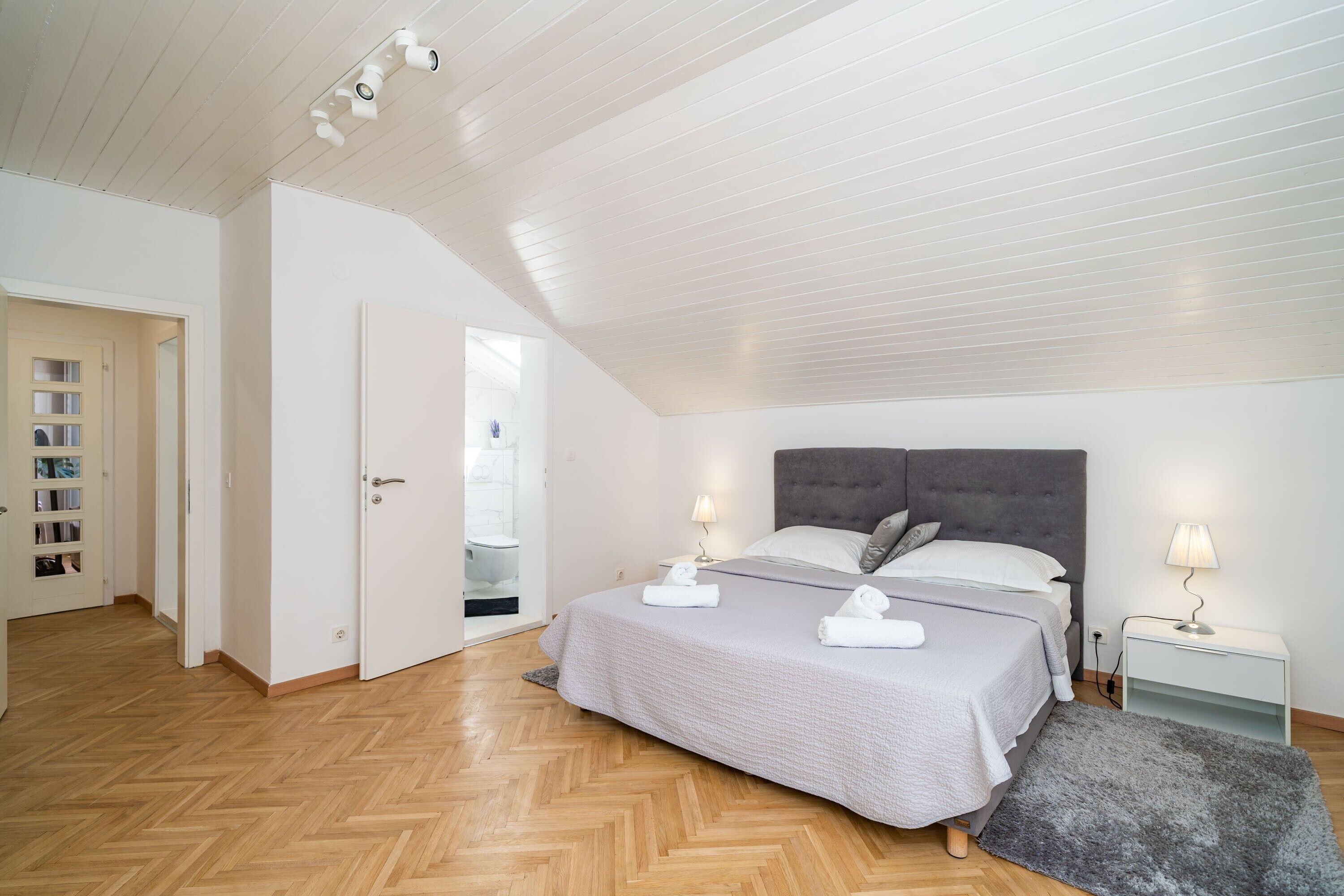 A bedroom with a bed, two bedside lamps, a grey rug, and a wooden herringbone floor.