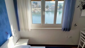 A hotel room with a bed, a window with a view of a body of water and buildings, a white bedside table, and a blue wall. - 3. Double room with sea view (Zadar)