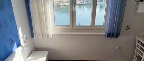 A hotel room with a bed, a window with a view of a body of water and buildings, a white bedside table, and a blue wall.