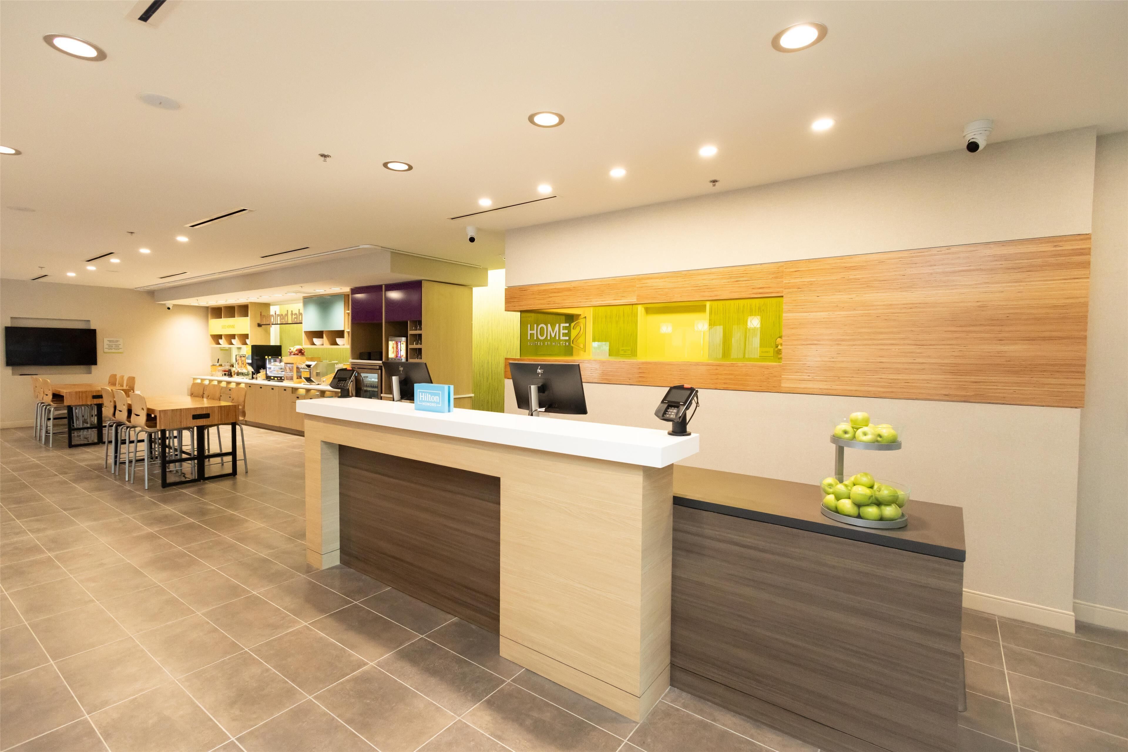 A hotel reception area with a reception desk, a seating area with tables and chairs, a television mounted on the wall, and a display of green apples on a stand.