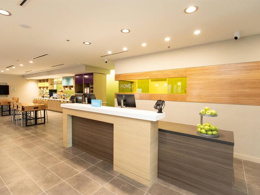 A hotel reception area with a reception desk, a seating area with tables and chairs, a television mounted on the wall, and a display of green apples on a stand.