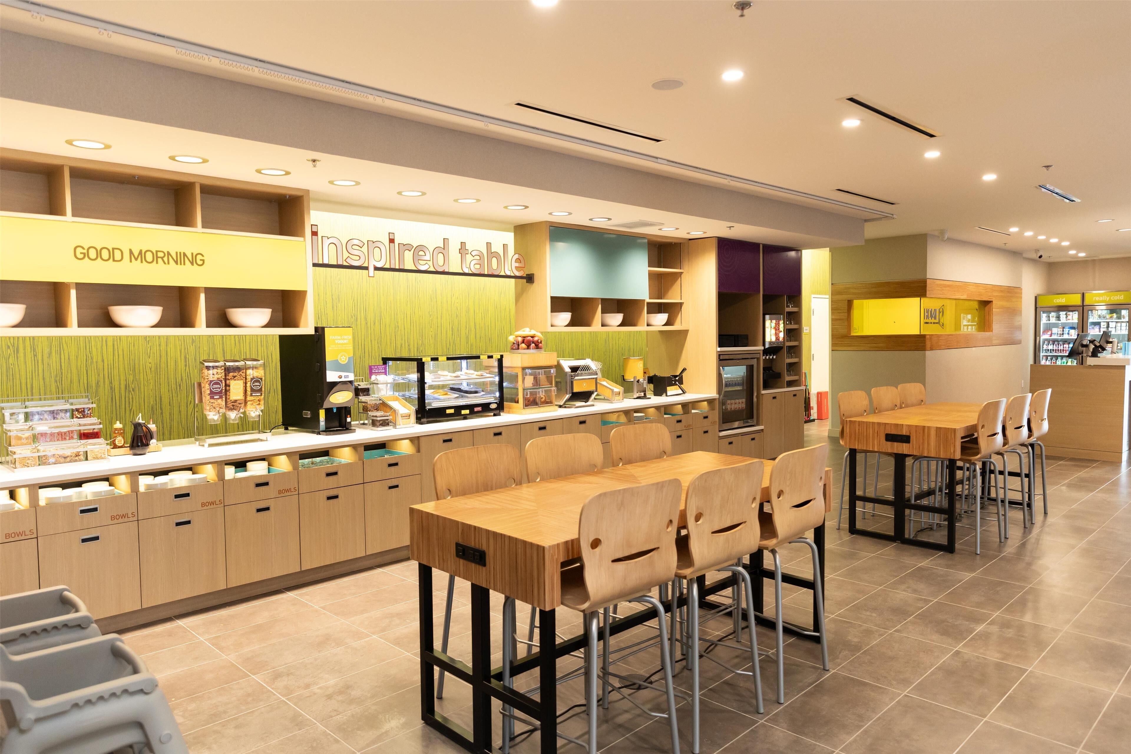 A modern cafe with a counter displaying various food items, a seating area with tables and chairs, and a 'Good Morning' sign above the counter.
