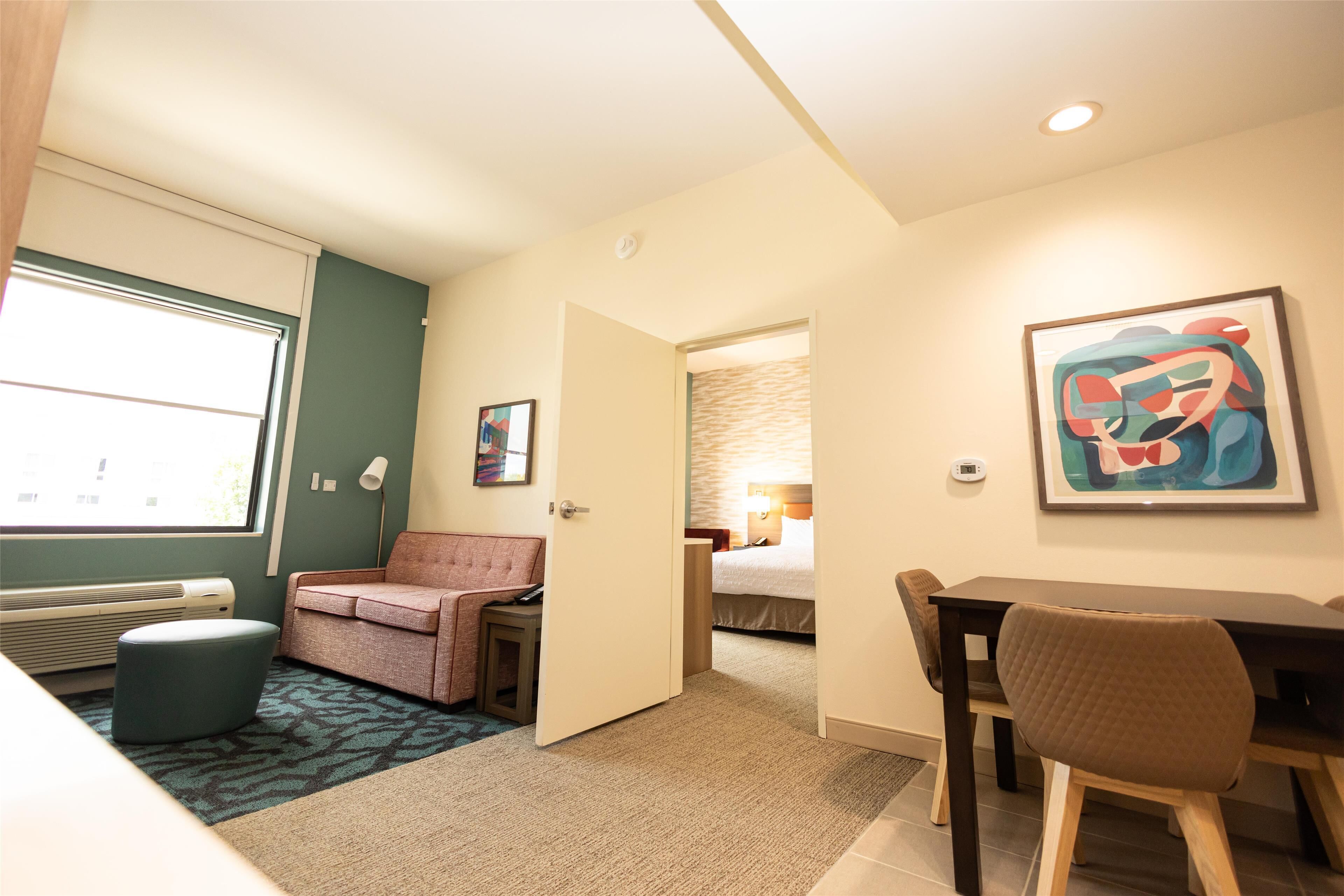 Une chambre d’hôtel compacte comprenant un lit, un bureau, une chaise, une petite table, un canapé, une fenêtre avec stores, un radiateur et un tableau au mur.