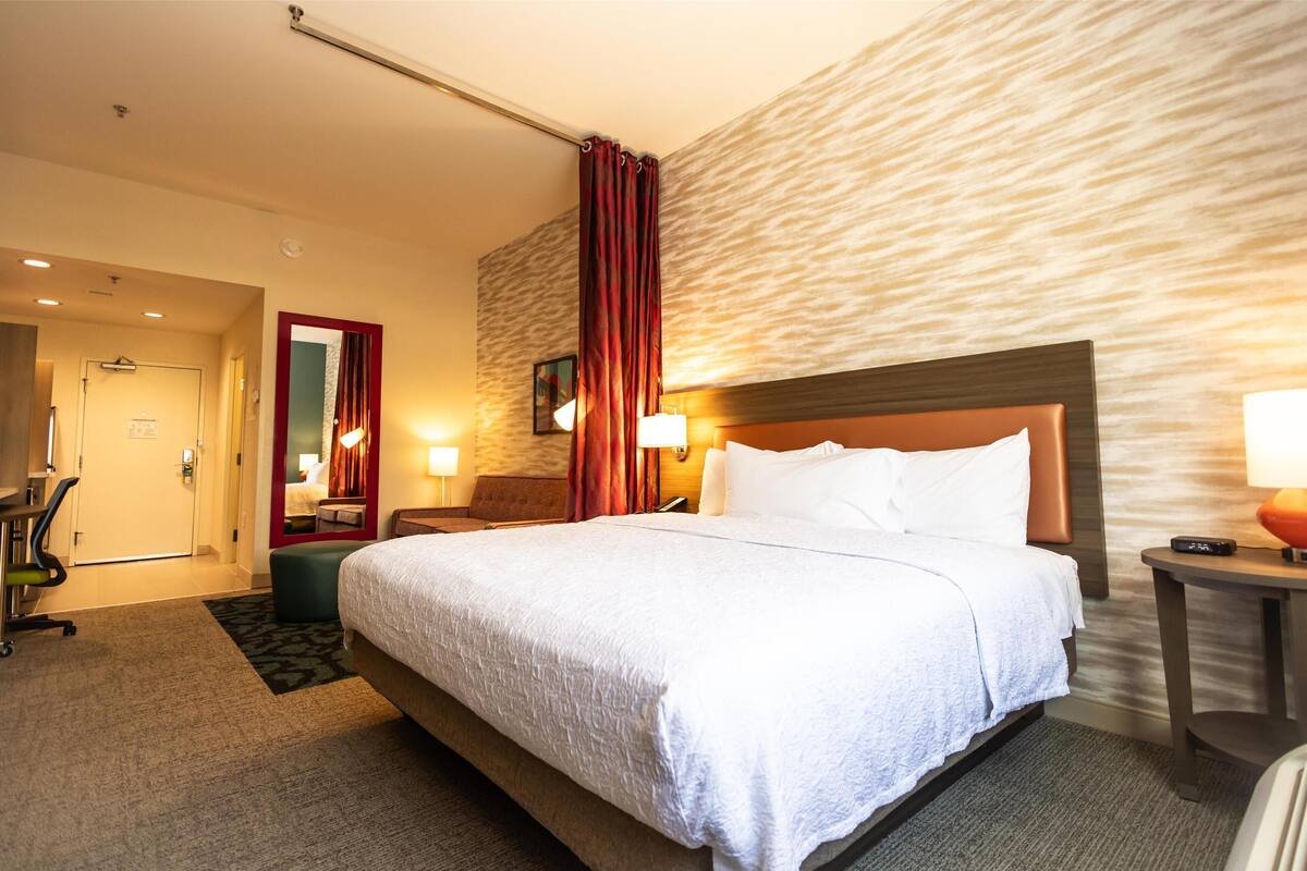 A hotel room with a large bed, a desk, a chair, and a mirror.