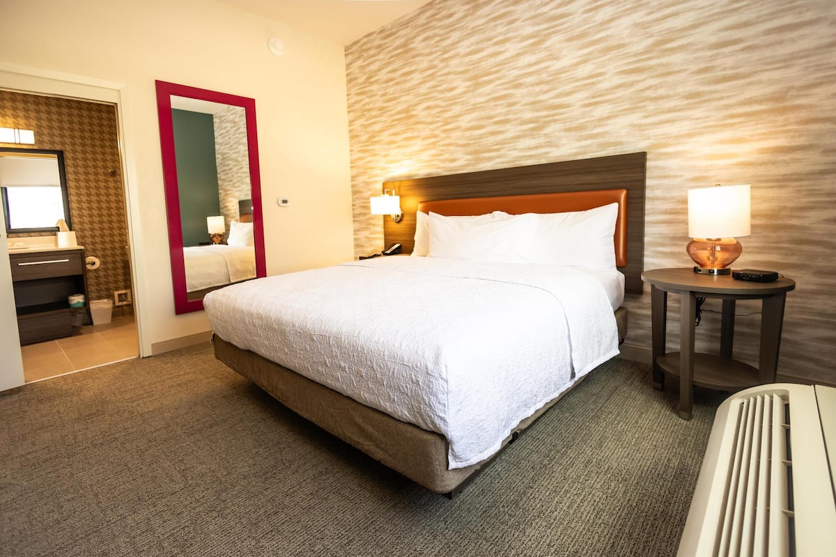 A hotel room with a large bed, a wooden headboard, a nightstand with a lamp, and a bathroom with a mirror and towel rack.