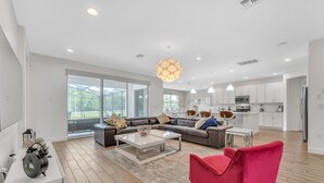 Ein modernes Wohnzimmer mit einem großen Ecksofa, einem roten Sessel und einem Couchtisch. Im Hintergrund ist eine Küche mit weißen Schränken und einer Mikrowelle zu sehen.