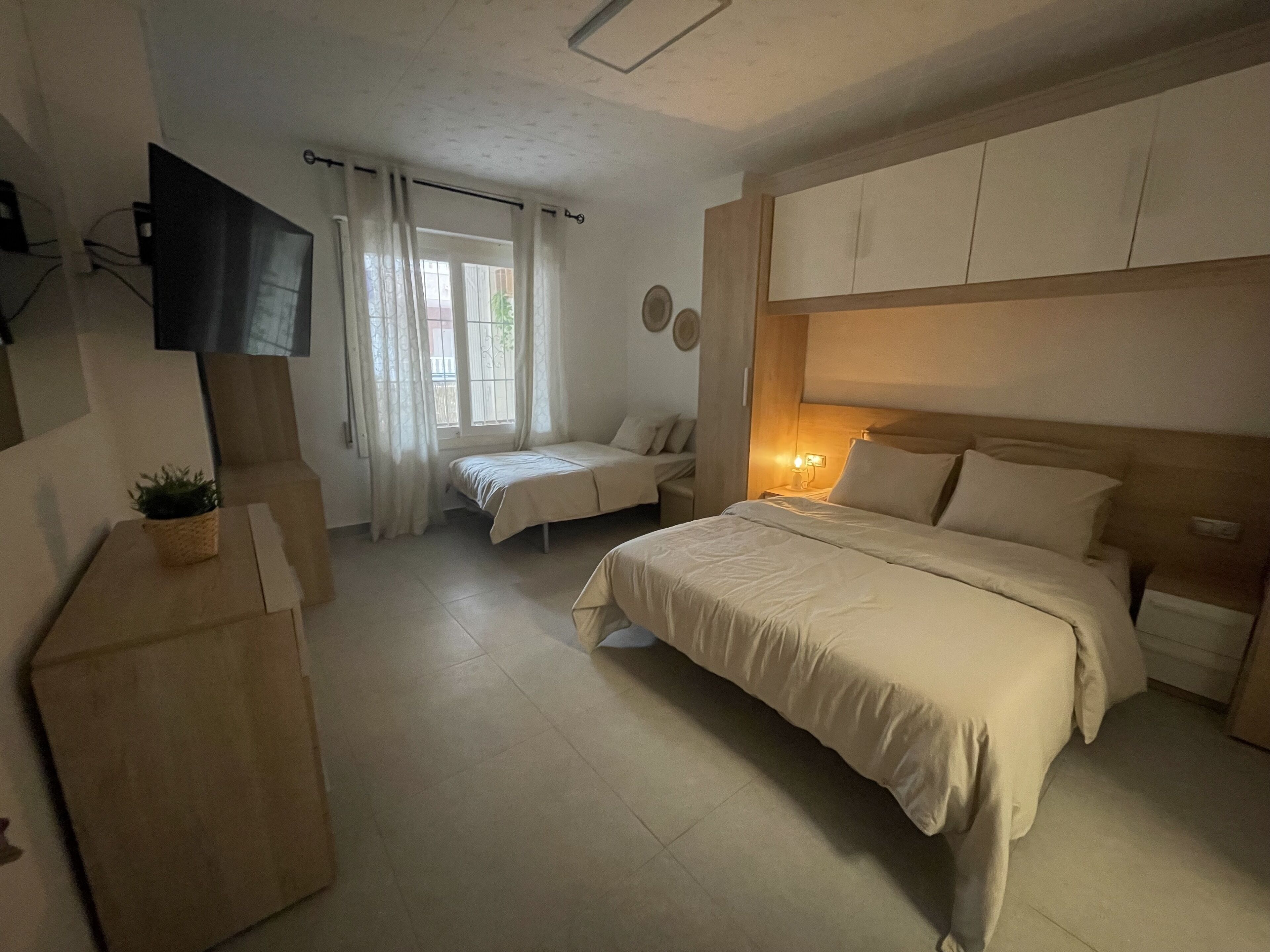 A bedroom with a bed, a bedside table, a TV mounted on the wall, and a window with curtains.