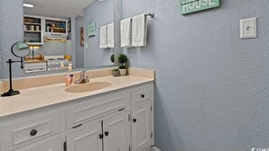 A bathroom with a large mirror, white cabinets, a beige countertop, and a sign that reads "BEACH HOUSE".