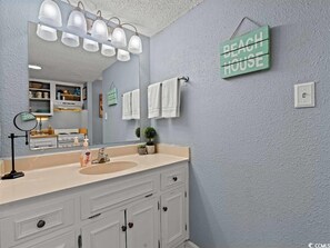 Un baño con un espejo grande, muebles blancos, encimera color beige y un cartel que dice "BEACH HOUSE".