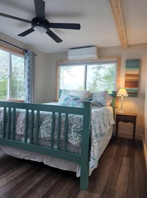 Cozy bedroom with a single bed, ceiling fan, and air conditioning unit, featuring a large window and wooden floor.