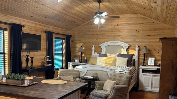 A cozy cabin-style room with a wooden ceiling, a bed with a white headboard, a striped sofa, a small dining table, and a kitchen area with a wooden countertop. - Cope's Haven (Fredericksburg)