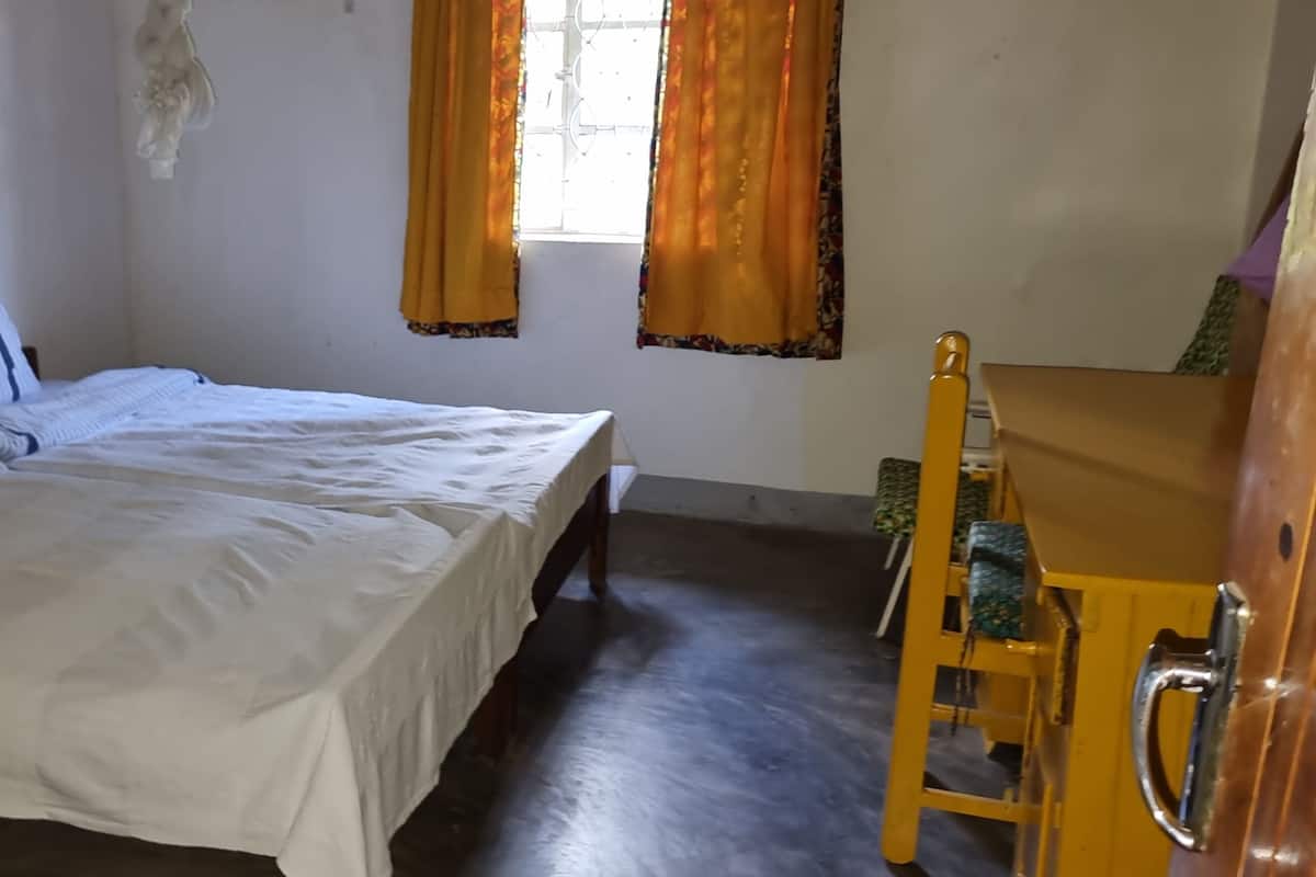 Ein Einzelbett mit einem gestreiften Kopfkissen und einer weißen Bettdecke, ein Holzstuhl und ein Fenster mit orangenen Vorhängen.