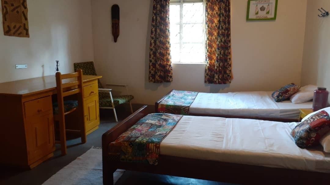 Una habitación con dos camas individuales, un aparador de madera, una silla y un ventanal con cortinas.
