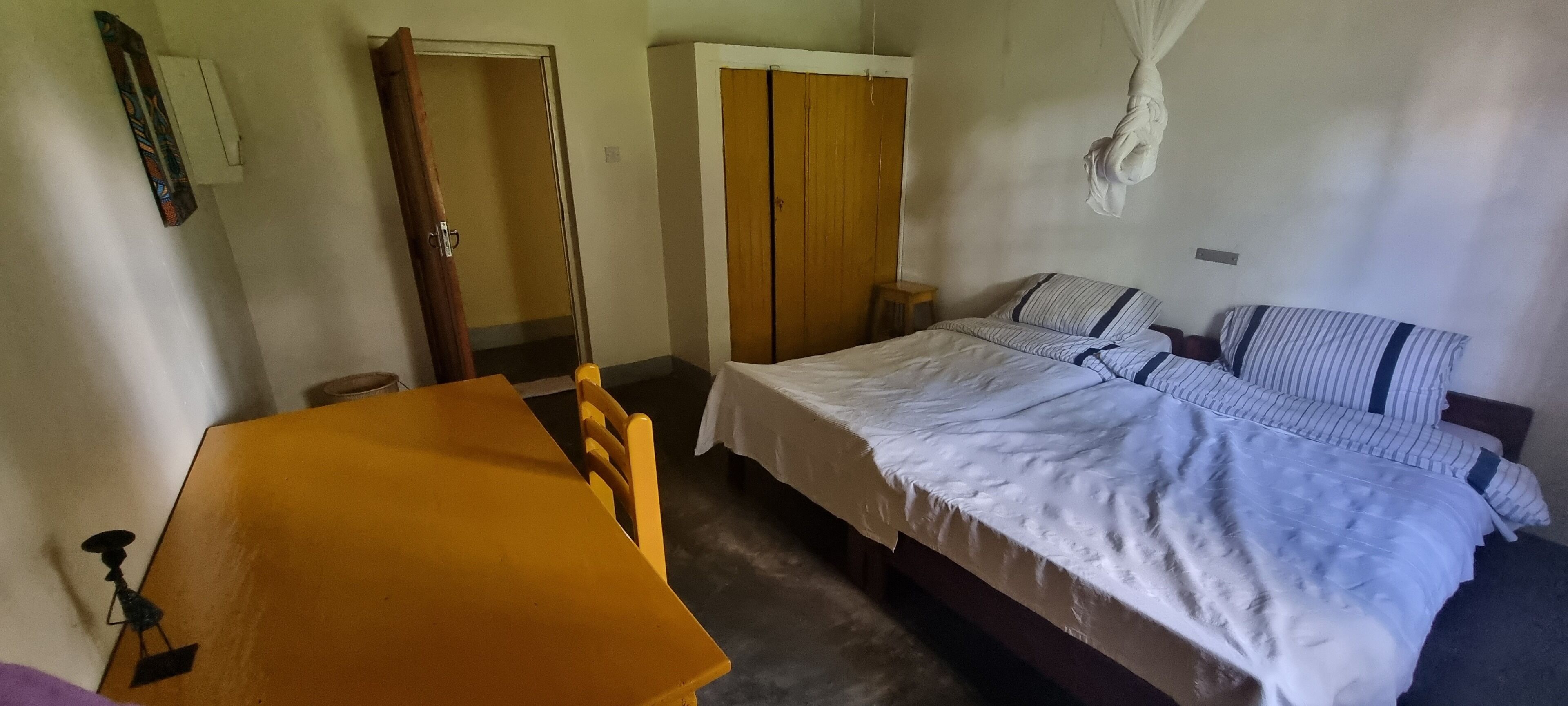 A bedroom with a bed, a wooden table, a chair, a wardrobe, and a hanging mosquito net.