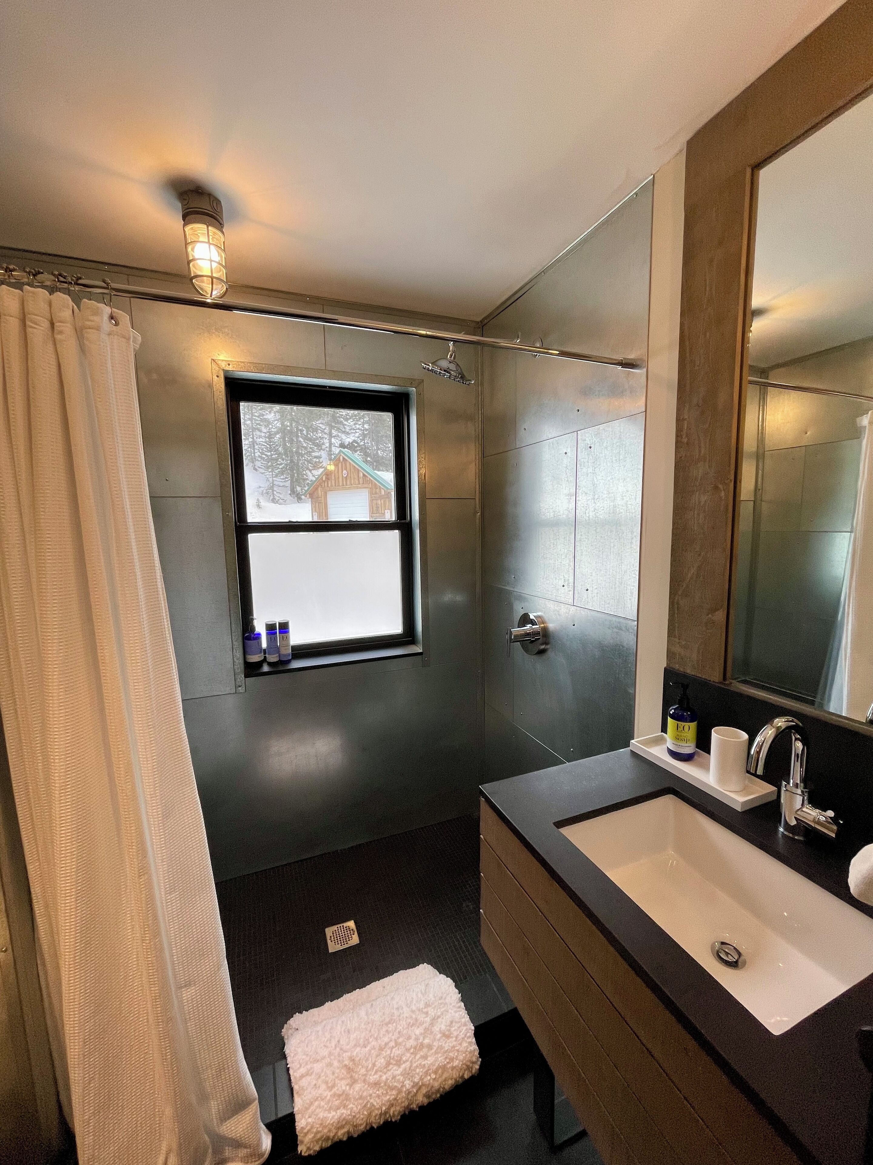 A modern bathroom with a large mirror, a sink, and a window with a view of a cabin.