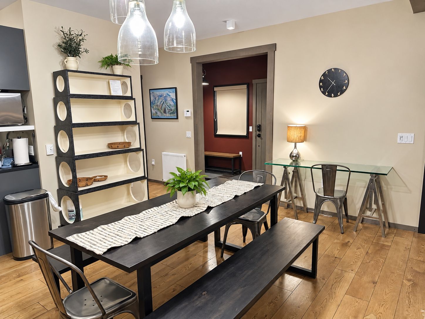 A dining area with a dark wooden table, benches, and chairs. There is a glass table lamp, a potted plant, and a wall clock.