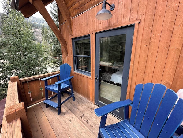 Une terrasse en bois avec deux chaises Adirondack bleues, une porte vitrée et une vue sur une zone boisée.