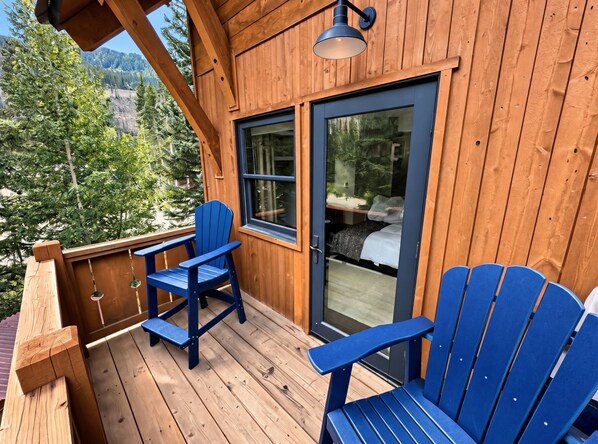 A wooden deck with two blue Adirondack chairs, a glass door leading to a bedroom, and a view of a forested area.