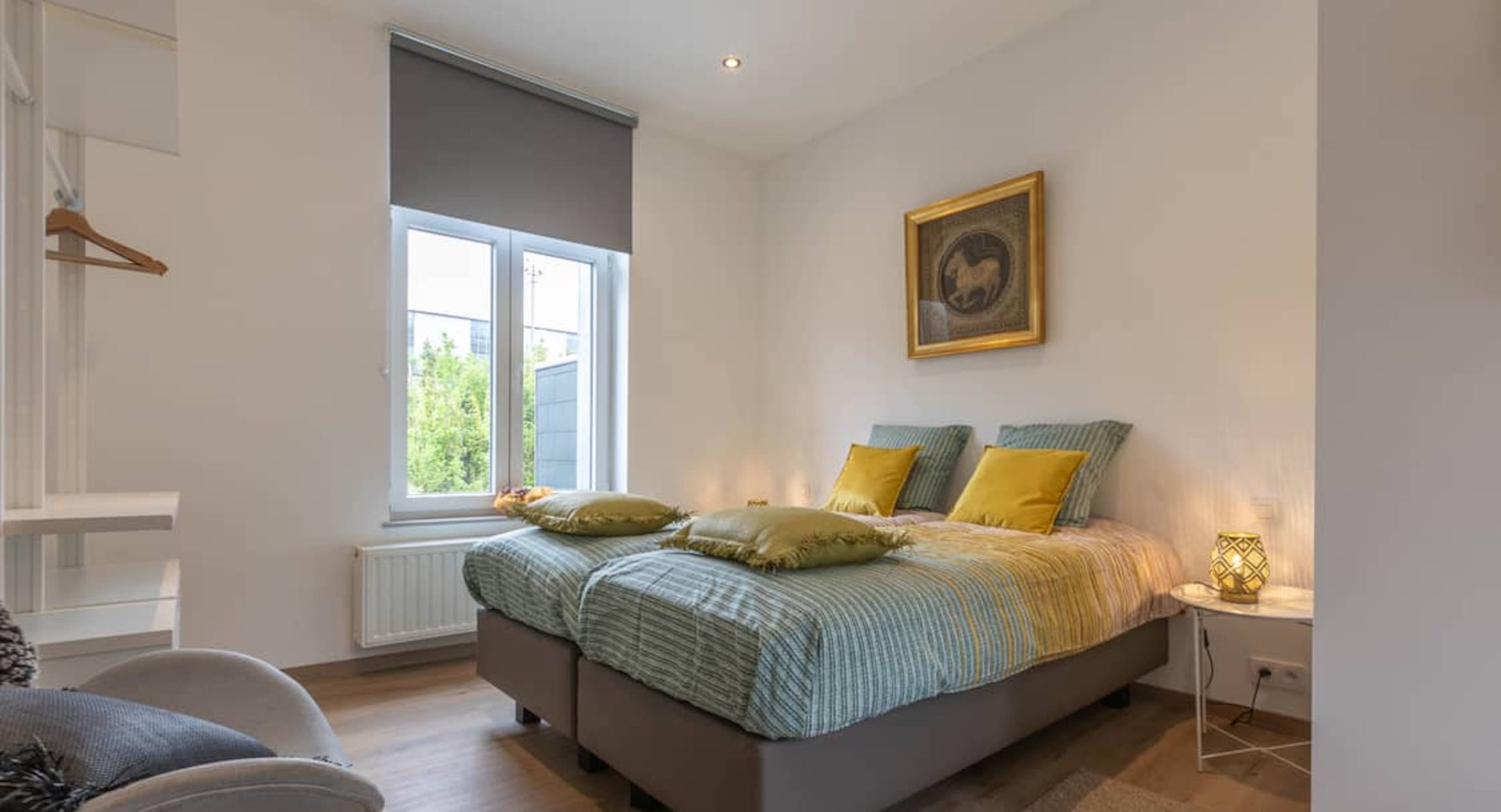 A bedroom with a bed, a window, a radiator, a chair, and a bedside table.