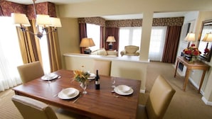 Un comedor de hotel con una mesa de madera dispuesta para cuatro personas, rodeada de sillas color beige. Hay dos lámparas sobre la mesa, un jarrón con flores y una botella de vino. La habitación tiene suelo alfombrado y vistas a un pasillo con un espejo y una pequeña mesa con un jarrón de flores.