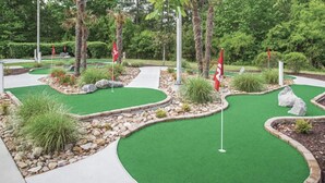 Un campo de mini golf con césped artificial verde, banderas rojas y zonas ajardinadas con rocas y plantas.