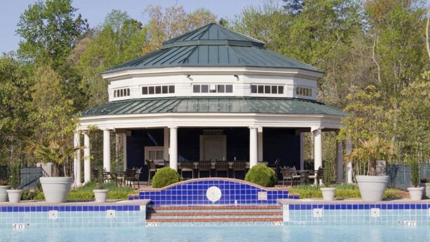 Un edificio de dos pisos con techo verde, un patio cubierto con asientos exteriores y una piscina con azulejos azules.