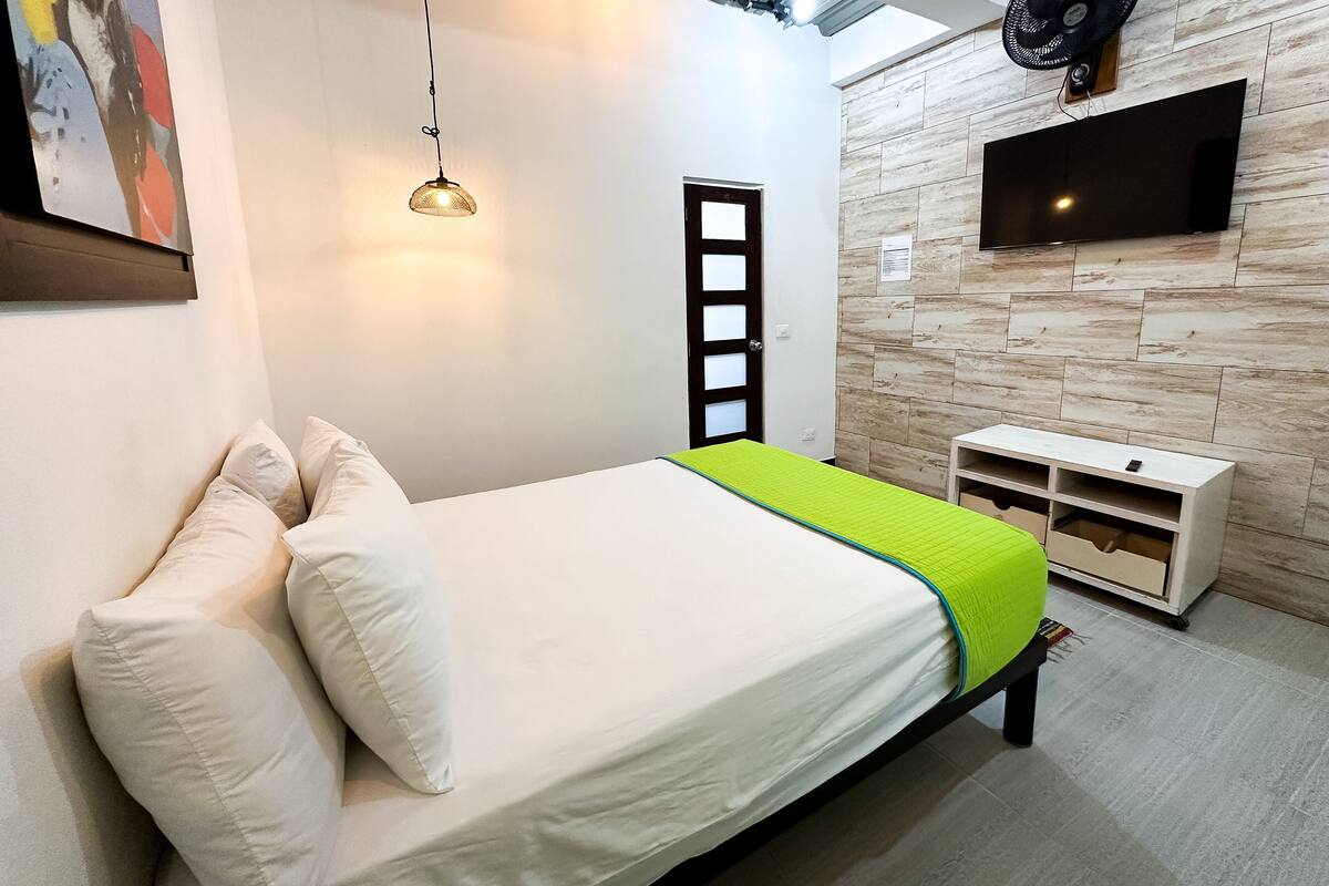 Une chambre à coucher avec un lit, des oreillers, une couverture verte, un téléviseur fixé au mur et une armoire blanche.