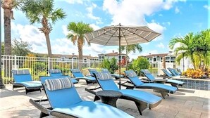 Zona junto a la piscina con sillas reclinables y sombrillas, rodeada de palmeras y una cerca blanca.