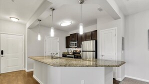 Una cocina moderna con encimeras de granito, electrodomésticos de acero inoxidable, iluminación colgante y armarios blancos.