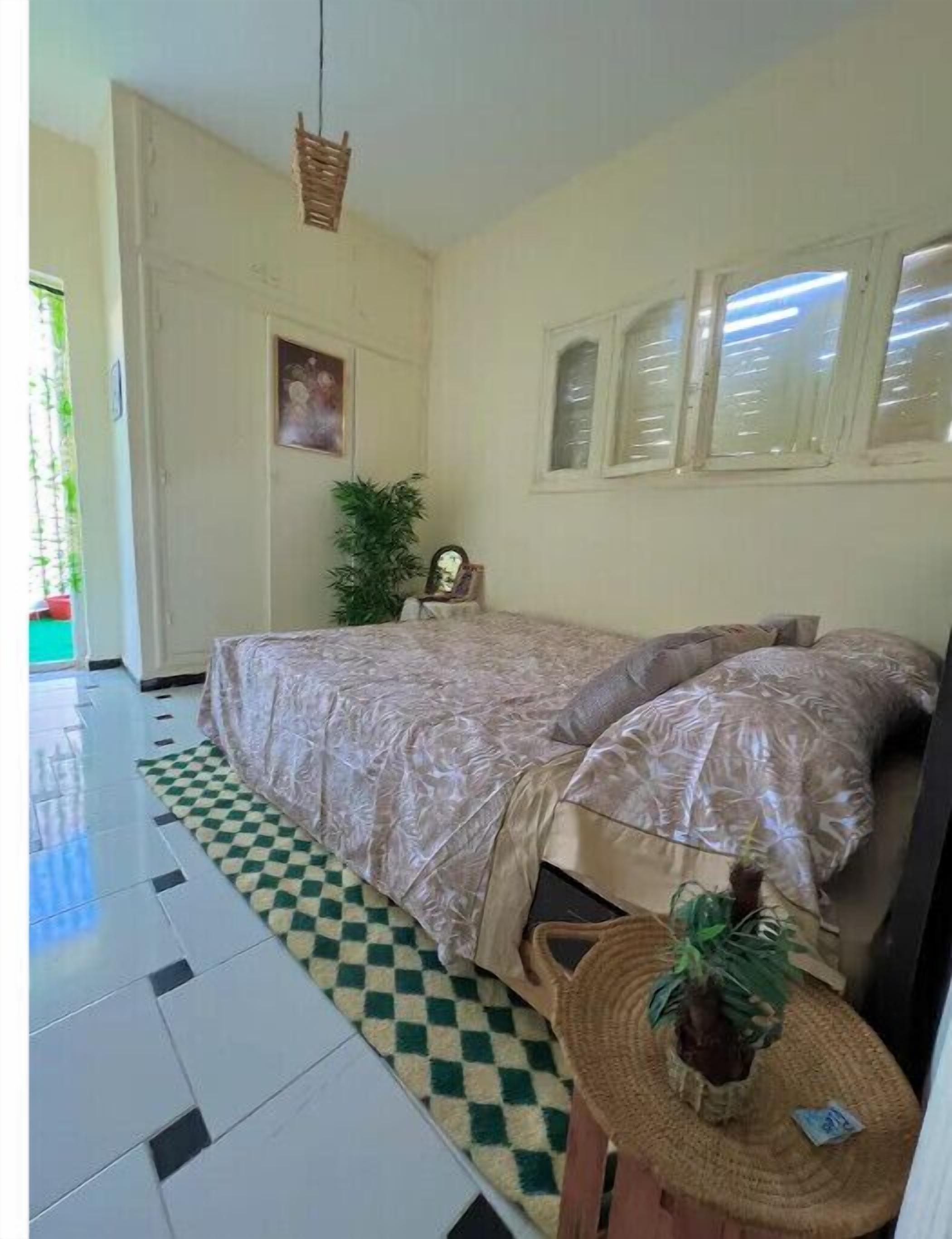 A bedroom with a bed, a checkered rug, a small round table with a plant, and a hanging light fixture.