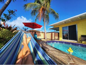 Un hamac avec vue sur une piscine, un parasol rouge et un bâtiment jaune.