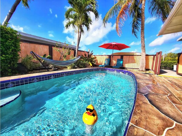 Une piscine avec un canard jaune flottant, des palmiers, un hamac et un parasol rouge.