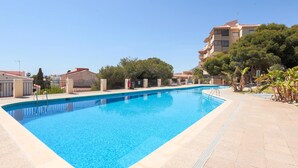 Une piscine rectangulaire à l’eau d’un bleu limpide, entourée d’une terrasse carrelée et d’une petite clôture. On aperçoit des arbres et un bâtiment au fond.