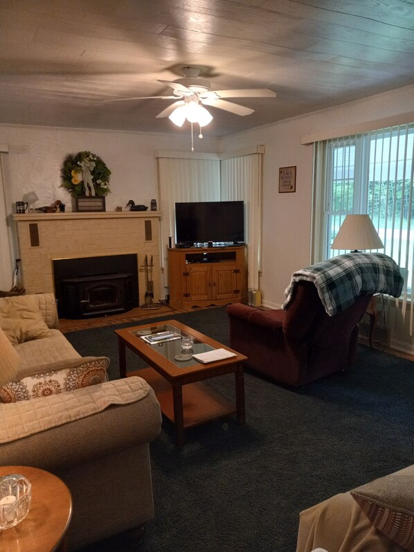 A living room with a fireplace, a television, a ceiling fan, and a window with blinds. - Cozy cottage across the street from Pymatuning lake (Pymatuning Central)