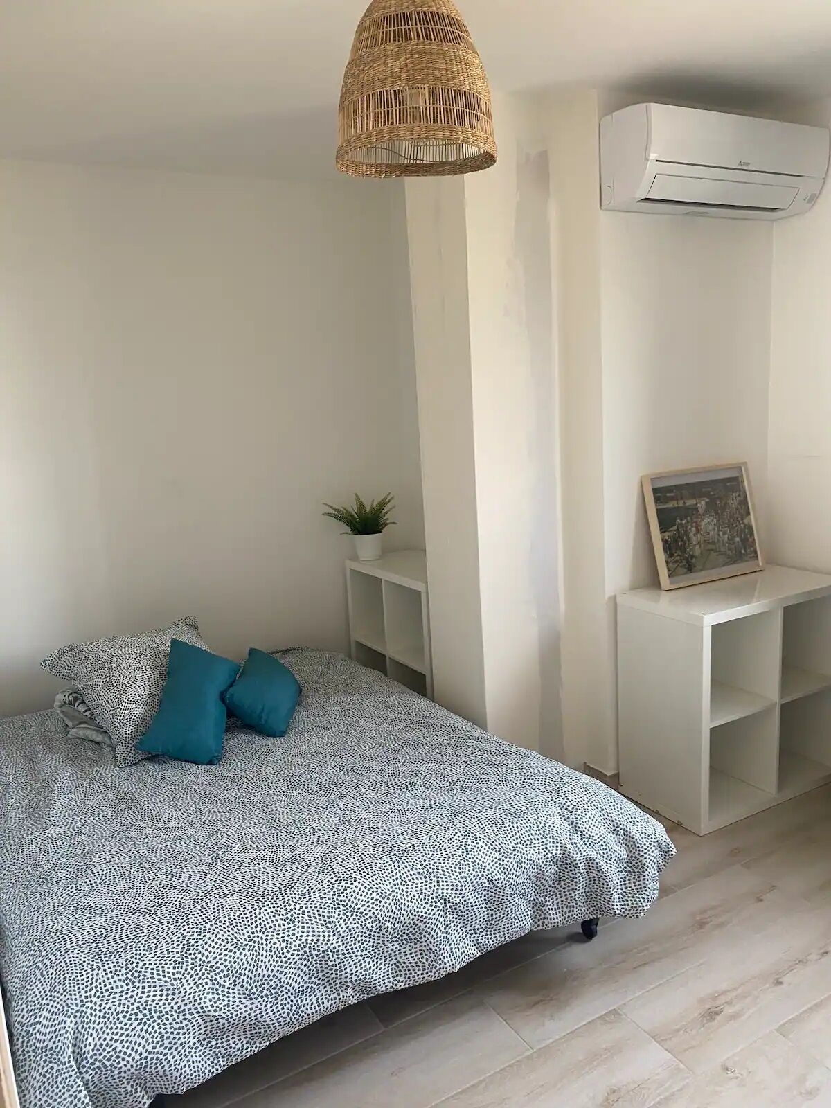 Une chambre à coucher avec un lit, un luminaire suspendu, un climatiseur, une petite plante et un cadre photo posé sur une console blanche.
