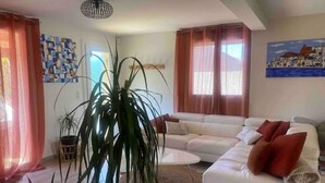 Una sala moderna con un sofá blanco, una mesa de centro y una planta grande. Hay cortinas en las ventanas y un cuadro en la pared.