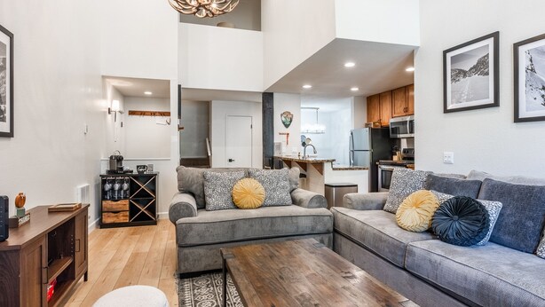 A modern living room with a sectional sofa, a coffee table, and a chandelier. There is a kitchen in the background with wooden cabinets and a refrigerator. - Bacta Haus 3br/3ba+Loft walk to Village & Canyon (Mammoth Lakes)
