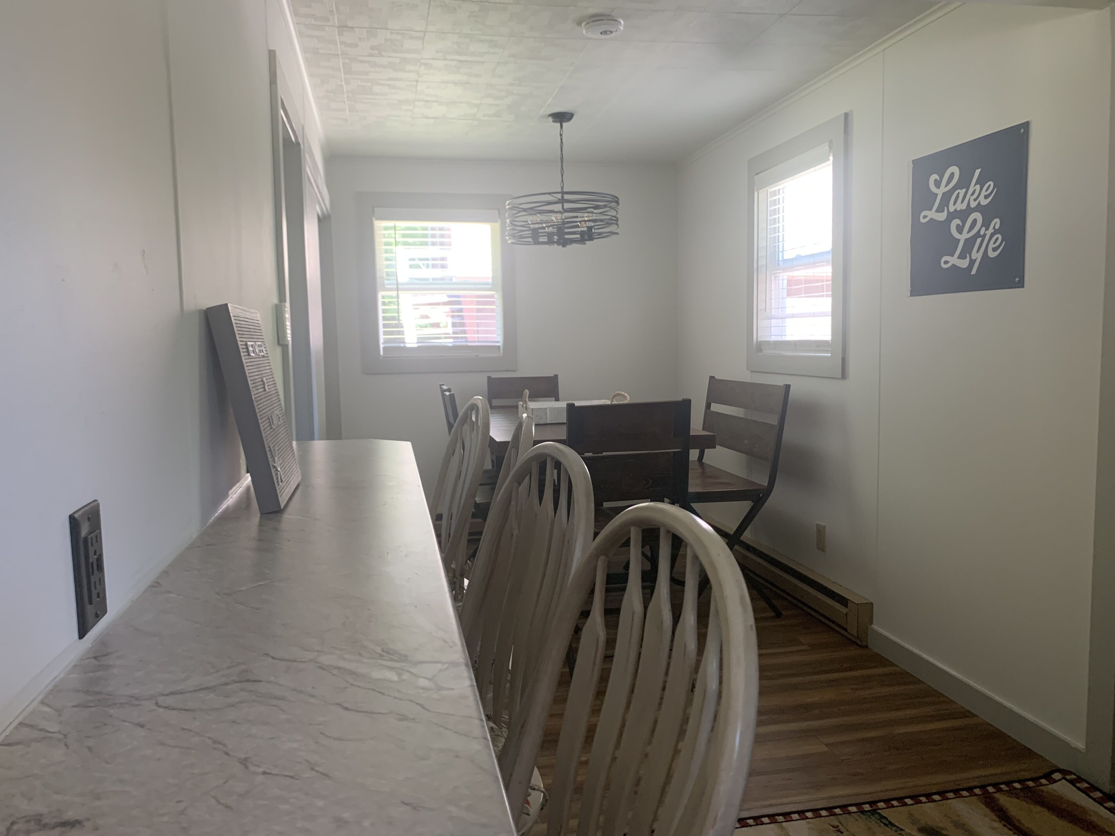 A dining area with a marble countertop, wooden chairs, a hanging light fixture, and a 'Lake Life' sign on the wall.
