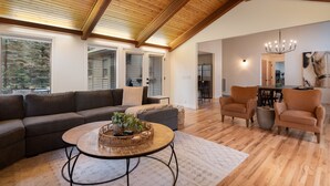 Ein geräumiges Wohnzimmer mit Holzboden, großen Fenstern, einer Couch, Sesseln, einem Essbereich und einem Kronleuchter.