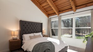 Ein Schlafzimmer mit einem großen Bett, einem Holzkopfteil, Nachttischlampen und einem Fenster mit Blick auf Bäume.