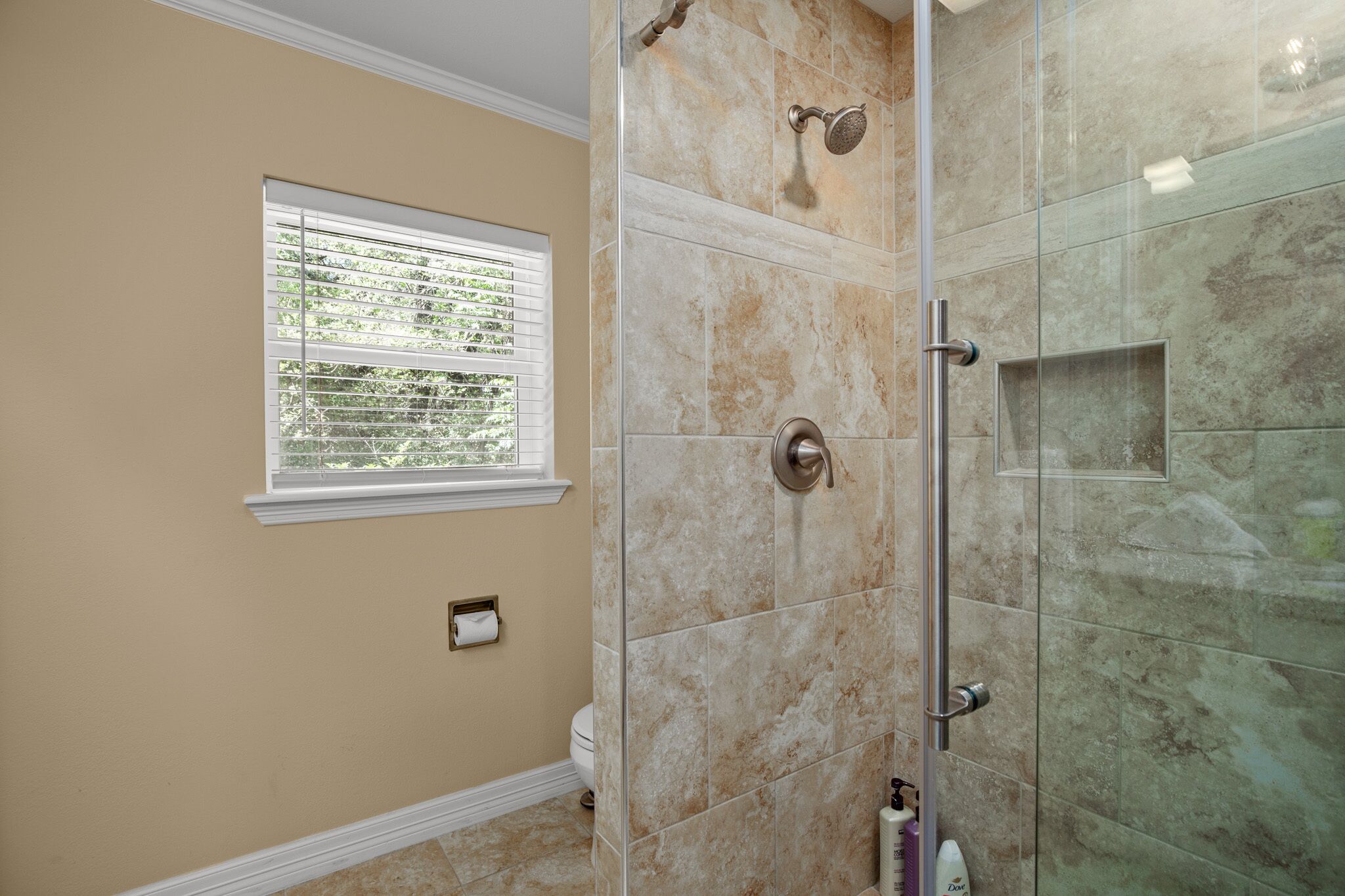 A bathroom with beige walls, a window with blinds, a toilet, and a glass-enclosed shower area.
