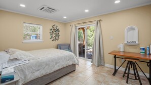 A bedroom with a bed, a desk, and a chair. There is a window with curtains, a wall-mounted air conditioner, and a small table with a tissue box and a can.