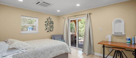 A bedroom with a bed, a desk, and a chair. There is a window with curtains, a wall-mounted air conditioner, and a small table with a tissue box and a can.