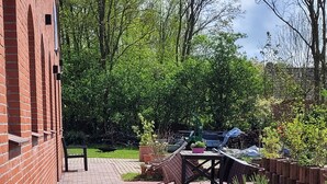 Ein gepflasterter Weg führt zu einer Terrasse mit Tisch und Stühlen, umgeben von Kübelpflanzen und einem Rasen.